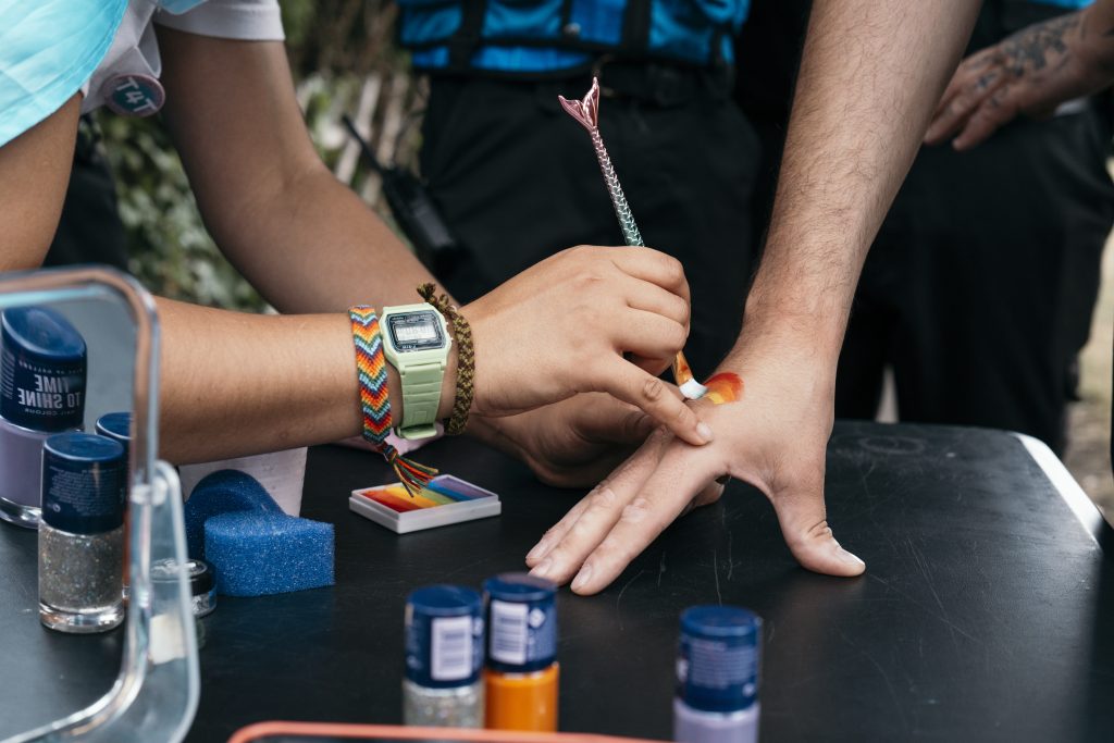 a person with a rainbow wristband painting a rainbow onto a security team member's hand using a mermaid tail brush