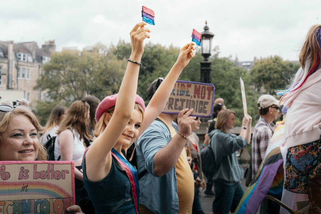 several people marching at Bath Pride 2025, including a person holding two tiny bisexual pride flags, a personal with a sign reading "life is better in technicolor" and a person with a sign reading "pride, not prejudice"