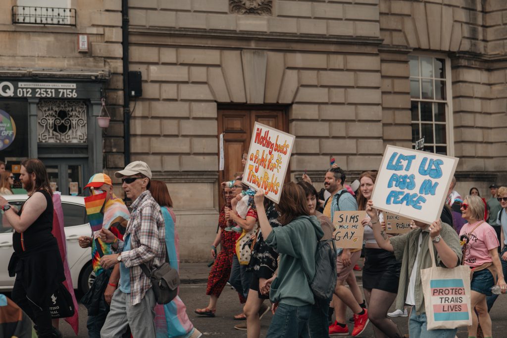 a group of people marching for Bath Pride 2025 holding several signs, including "Let us piss in peace", "Sims made me gay", "Nothing beats a fight for trans rights" and "protect trans lives"